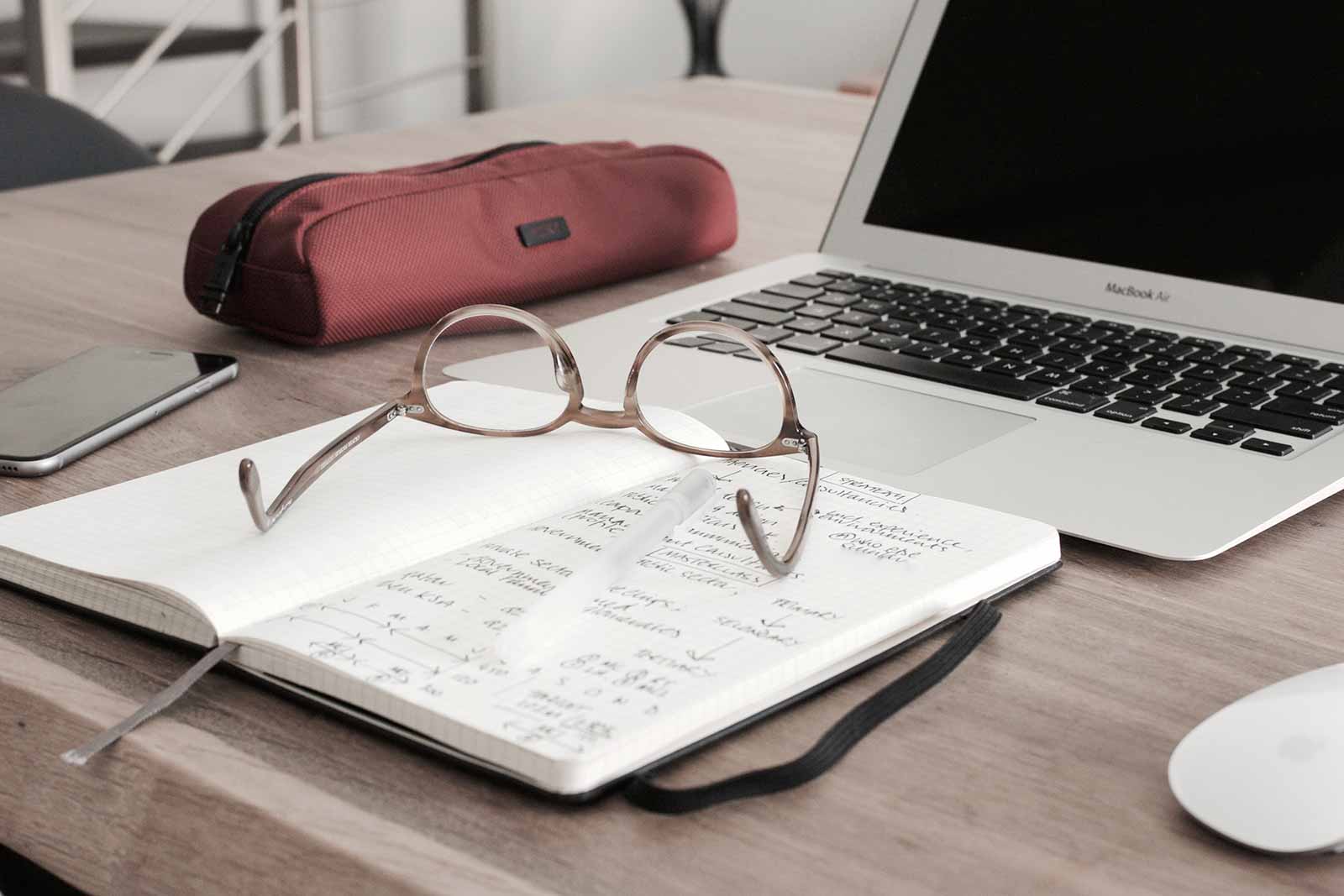 Workspace with glasses, notebook, and laptop on a wooden desk.