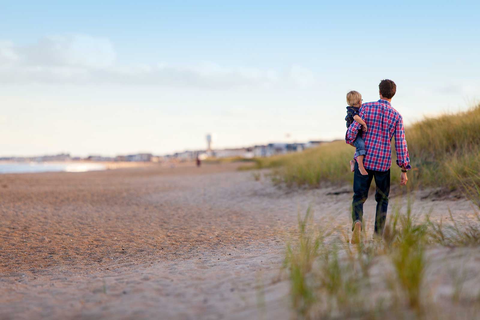 A father carrying his child on a sandy beach with a lighthouse in the distance.
