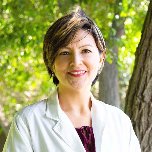 Smiling woman in a white lab coat outdoors with greenery.