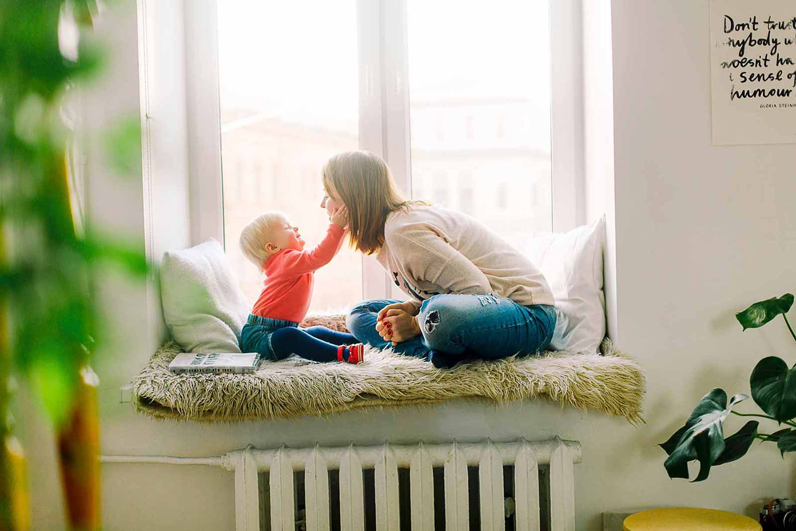 A mother lovingly kisses her child near a sunlit window.