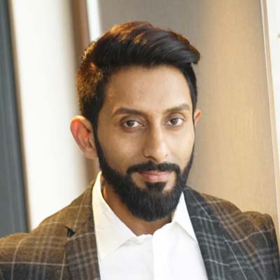 A man with a beard wearing a checked blazer and white shirt.